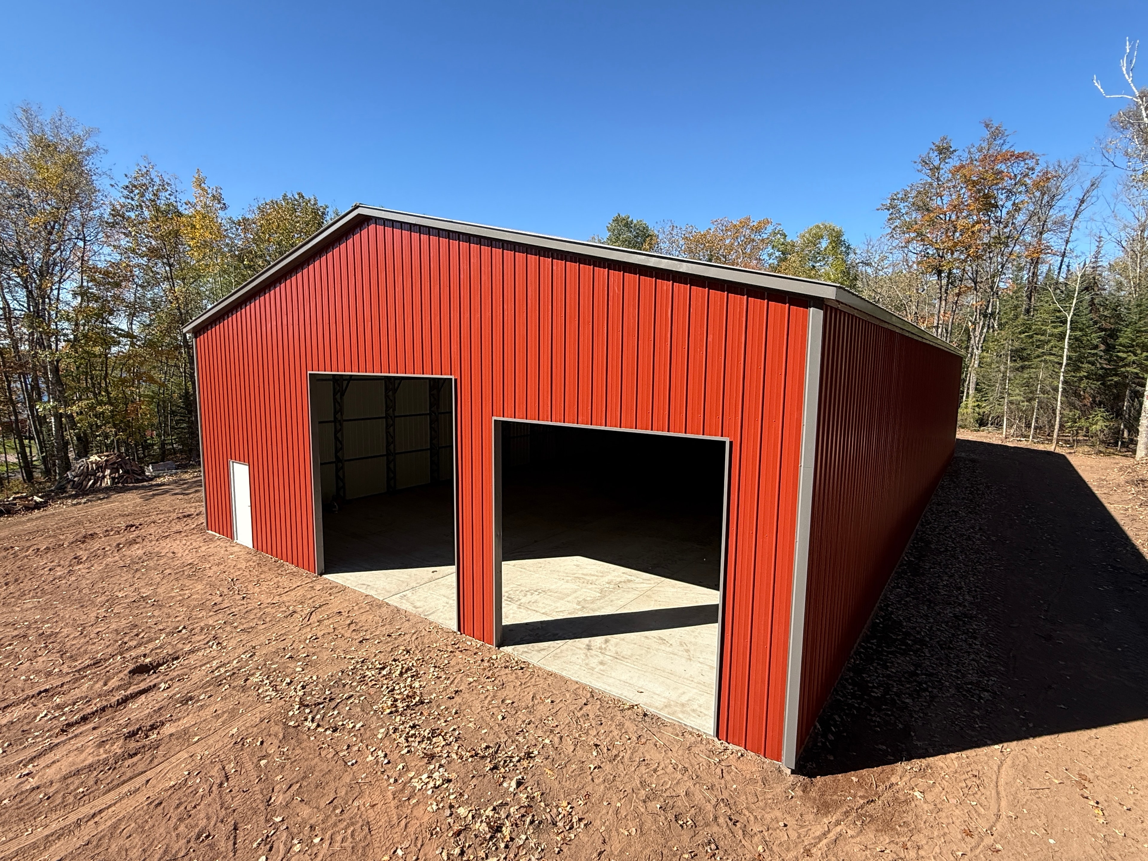Barn red metal garage with open roll-up door and walk-in door on a gravel and concrete pad in a wooded setting