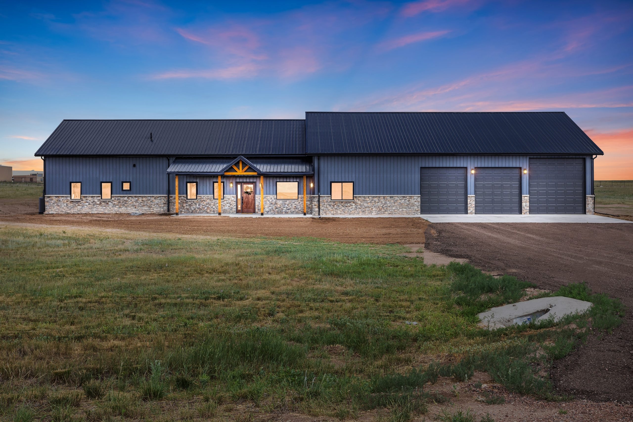 Charcoal metal barndominium at sunset with stone wainscot, wood entry accents, and attached garage