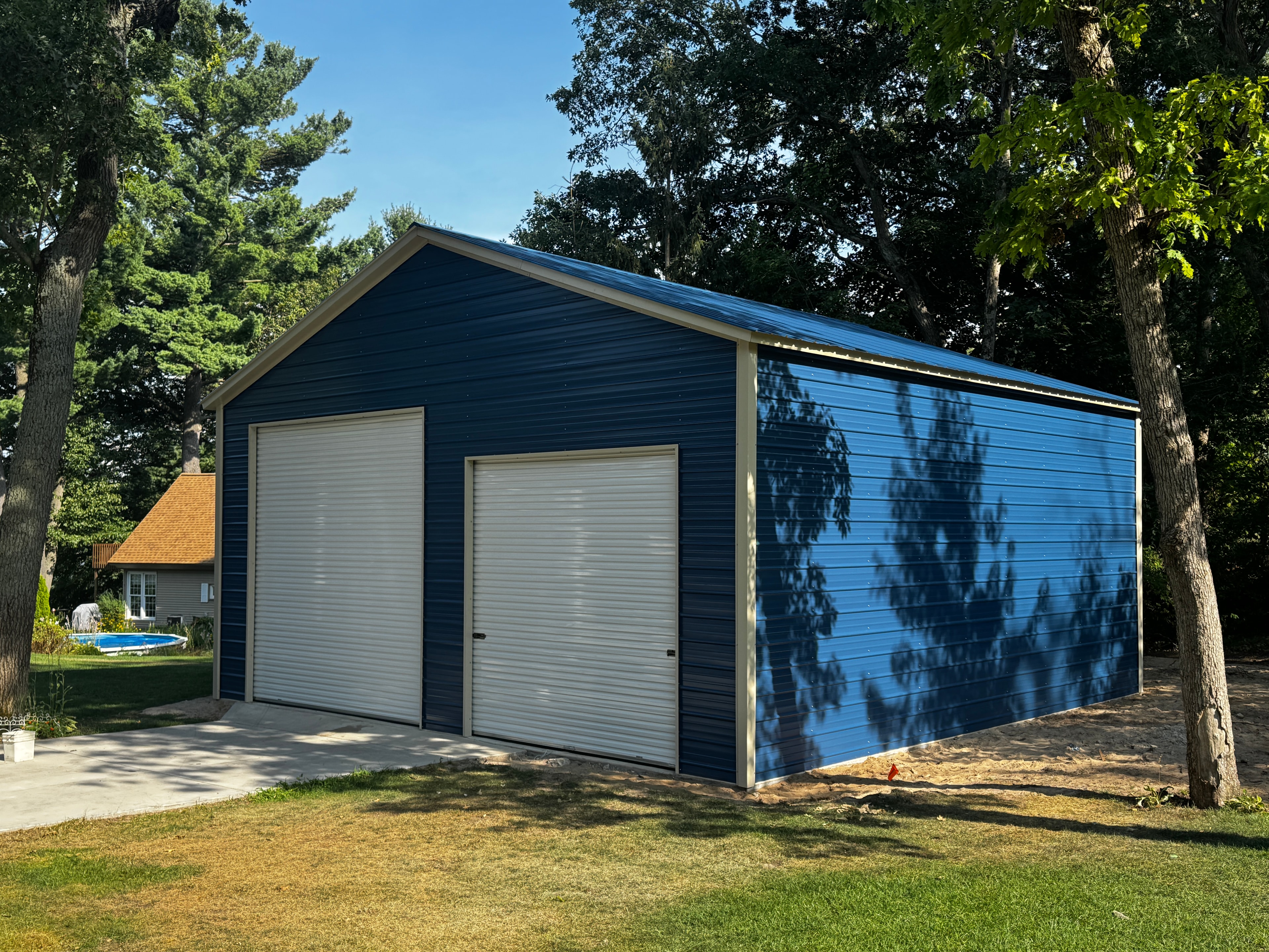 Blue metal garage with horizontal siding, two white roll-up doors, and walk-in door on a concrete pad