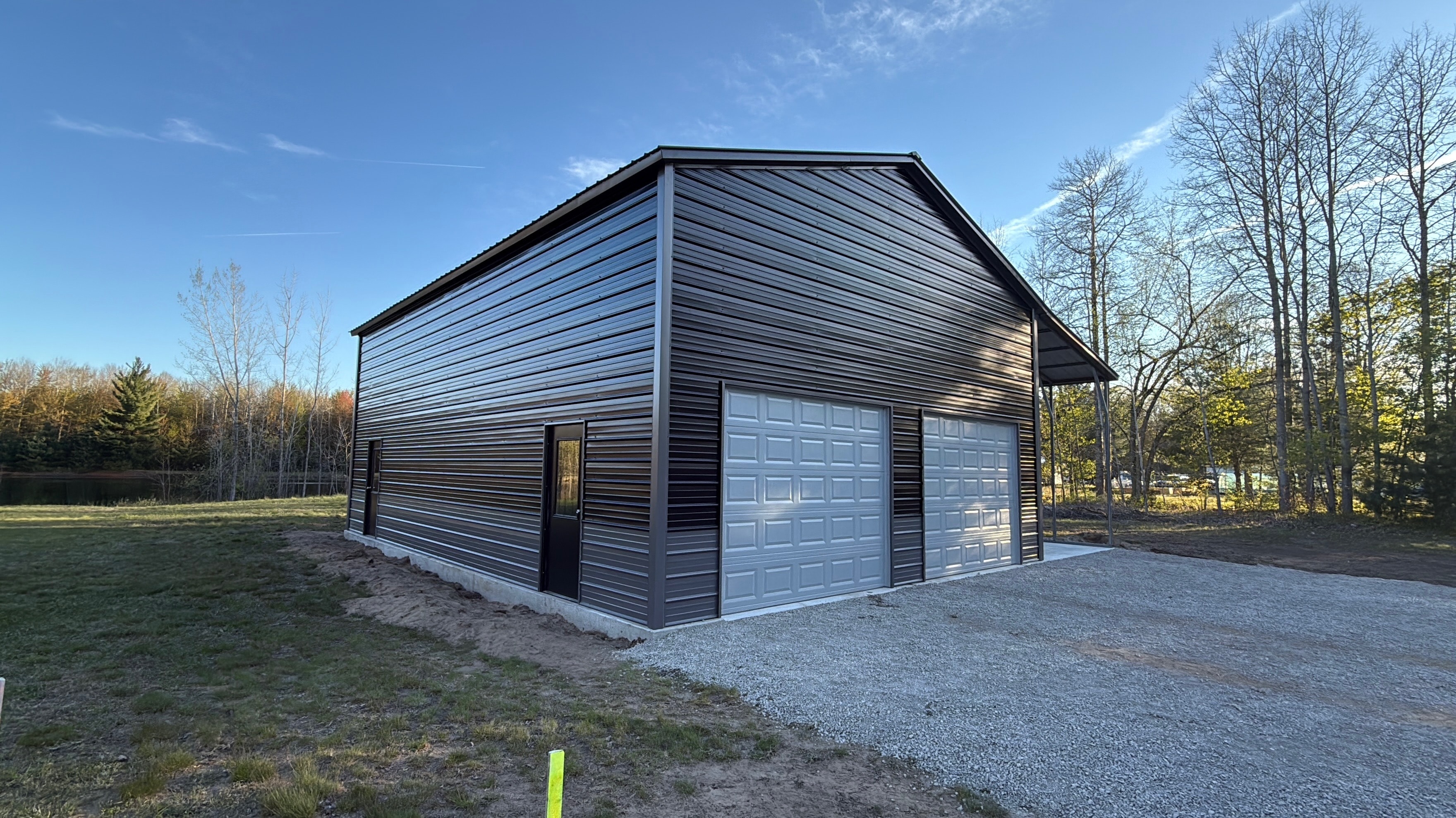 Charcoal metal garage with lean-to overhang, white roll-up door, walk-in door, and window on a gravel pad near a lake