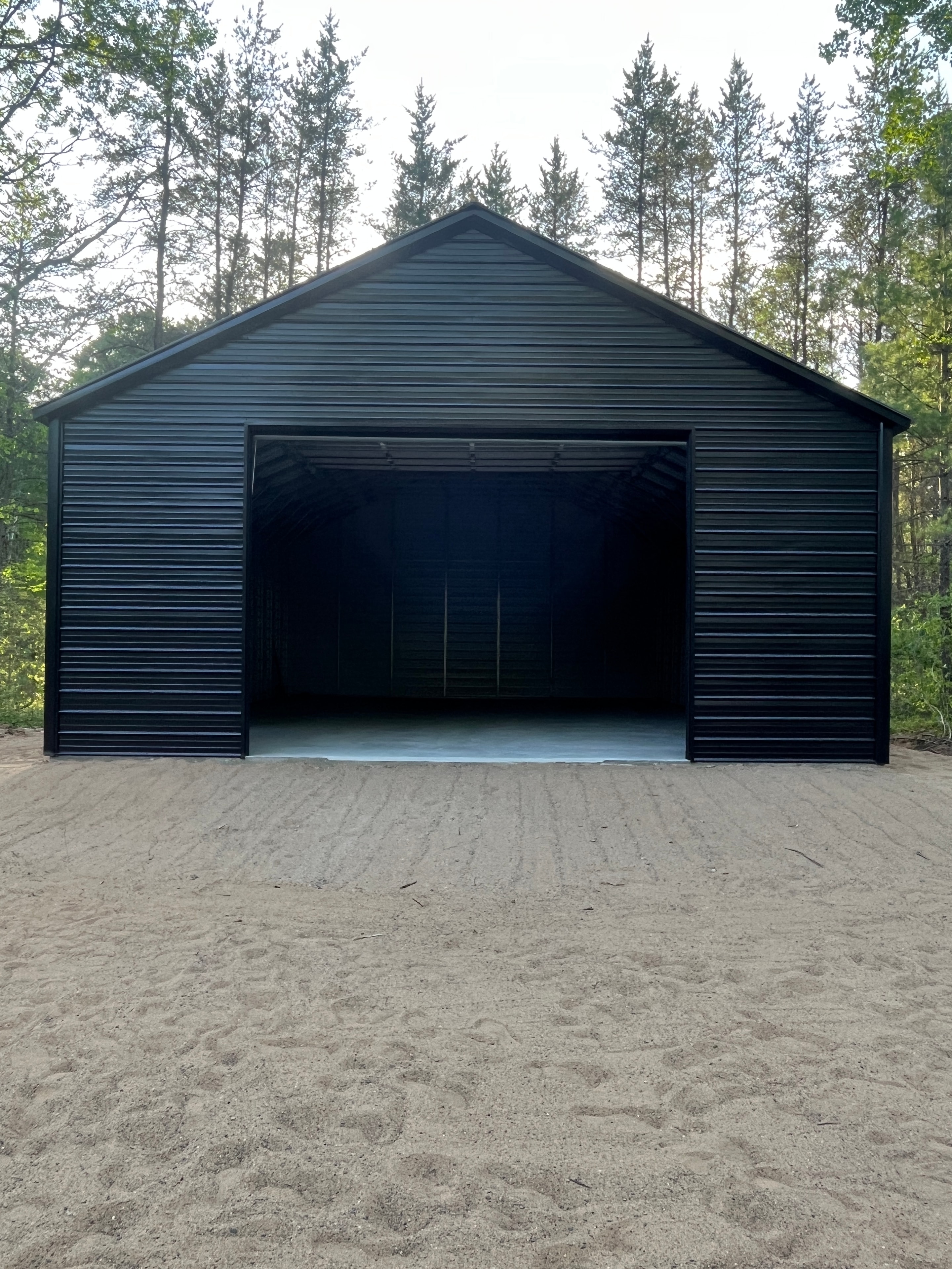 Charcoal metal open-front garage on a sandy lot surrounded by trees