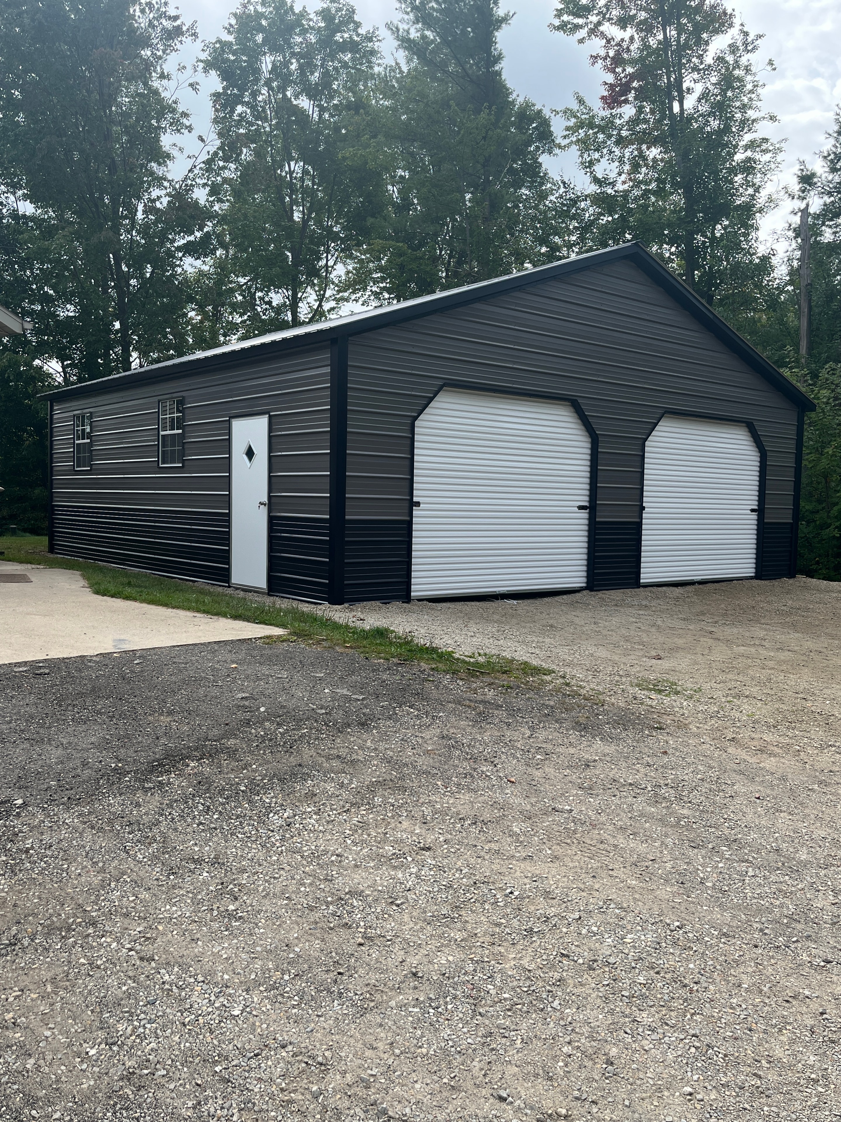 Charcoal metal workshop with three white roll-up doors, walk-in door, and window in a wooded setting