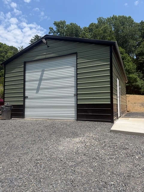 Green metal garage with dark brown trim and large white roll-up door on a gravel pad with wooded background