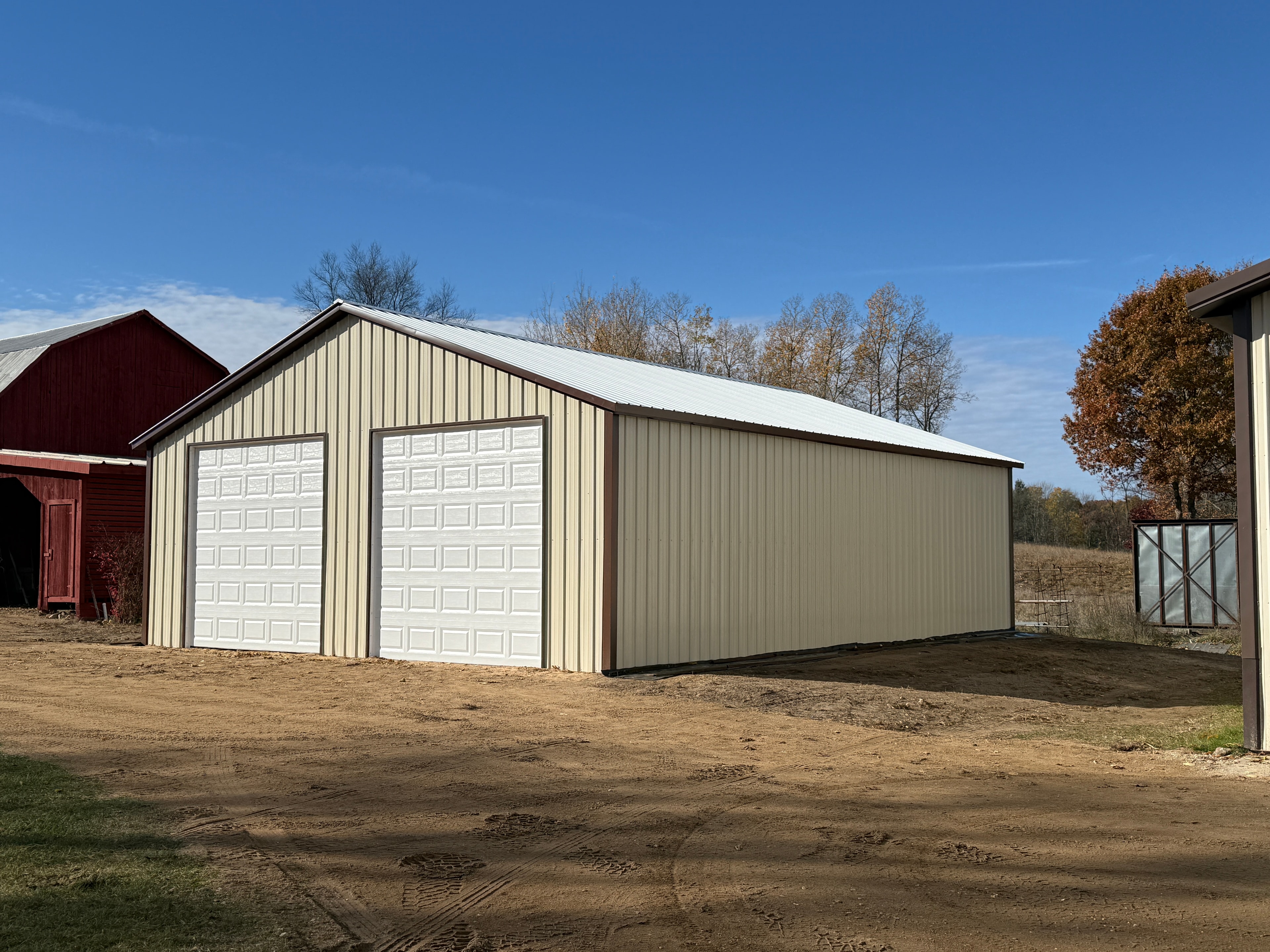 Side view of a tan metal workshop with brown trim, two white roll-up doors, and galvalume roof on a farm property