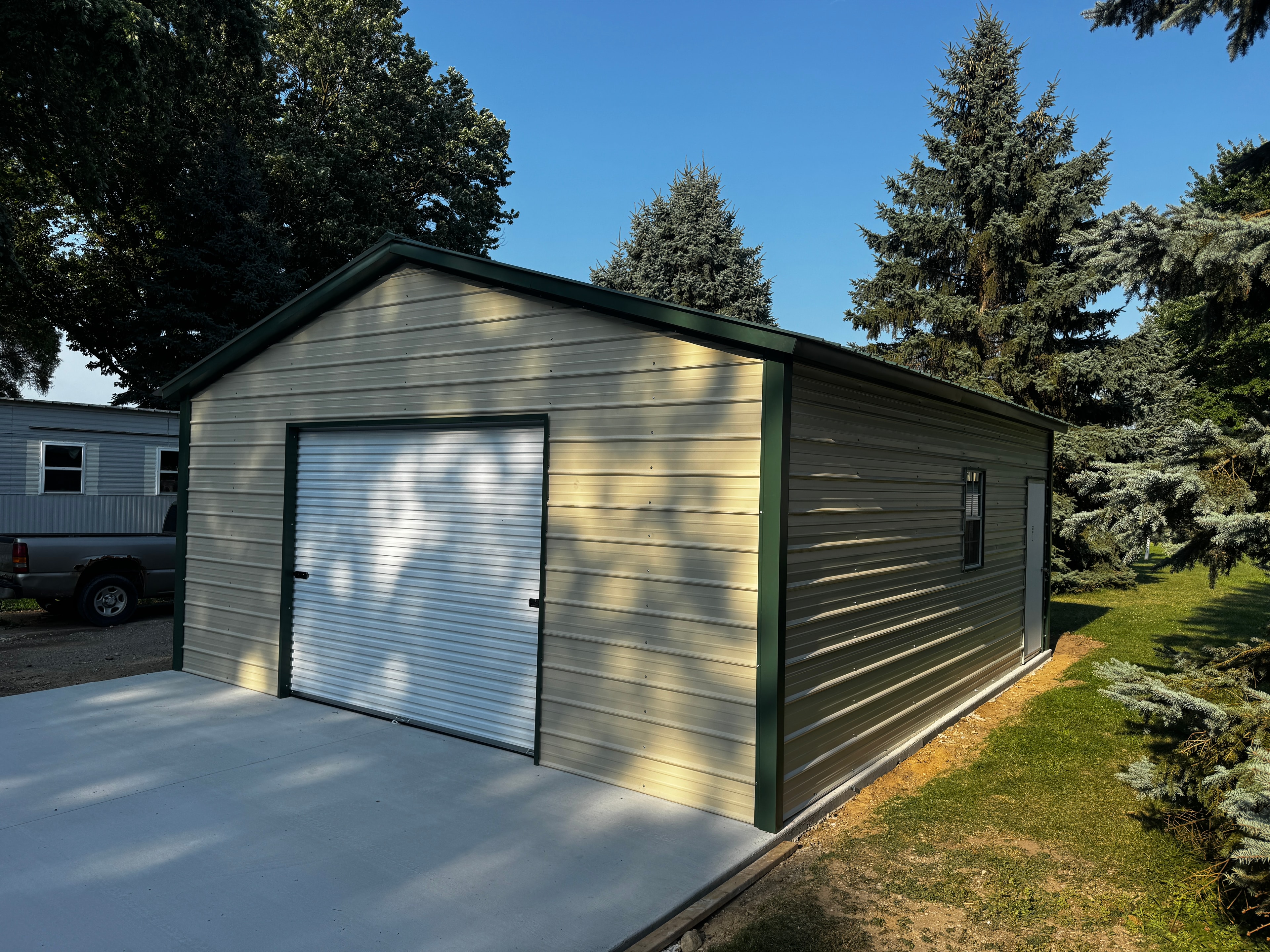 Tan metal garage with dark green trim, horizontal siding, one roll-up door and a window